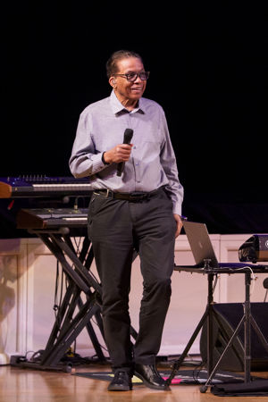 Herbie Hancock smiles on stage and holds a microphone, wearing a blue checkered button-up, gray pants and glasses