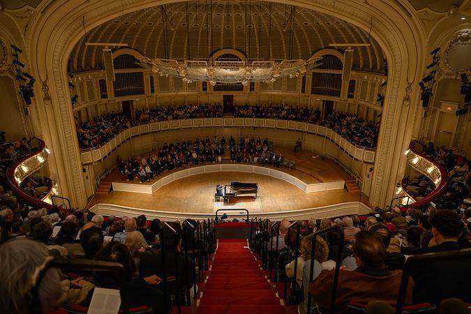 a wide shot from the balcony of pianist Yunchan Lim performing and audience members watching intently from both seats and stage