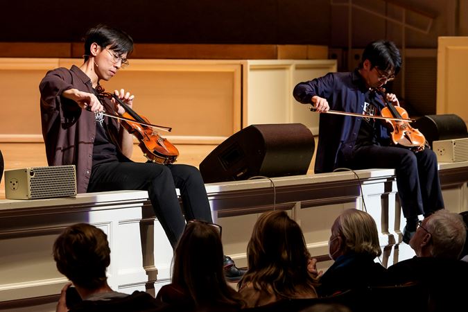 Eddy and Brett play their violins like cellos while sitting on the edge of the stage