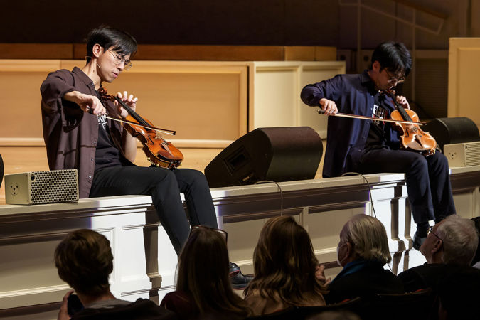 Eddy and Brett play their violins like cellos while sitting on the edge of the stage