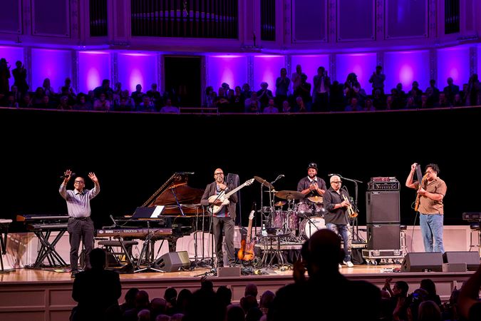 the full ensemble takes a bow on stage as the audience rises to its feet against the purple backlighting