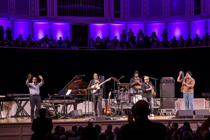the full ensemble takes a bow on stage as the audience rises to its feet against the purple backlighting