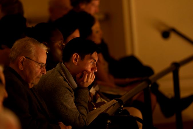 an audience member in the upper balcony with short dark hair is enraptured by the performance, chin resting in hand