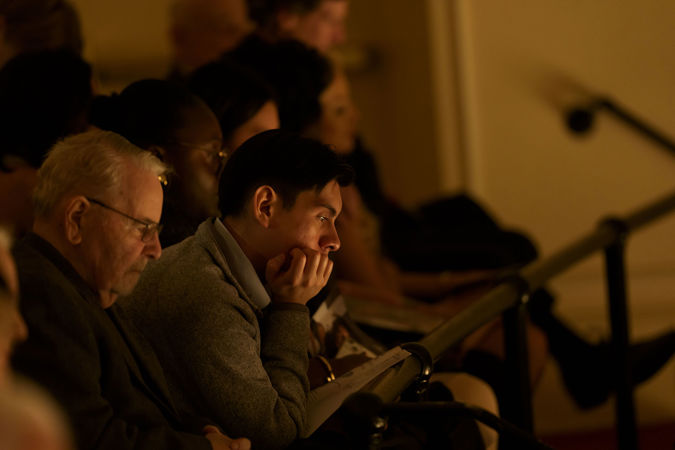 an audience member in the upper balcony with short dark hair is enraptured by the performance, chin resting in hand