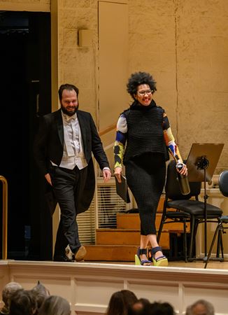 Julia Bullock, in a black knit outfit with colorful sleeves, walks out onstage, trailed by conductor Petr Popelka