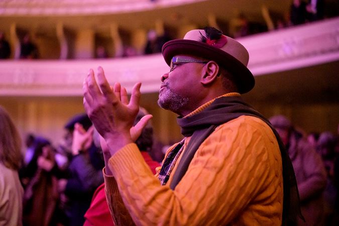an audience member in a brown hat and orange sweater stands and claps after the performance