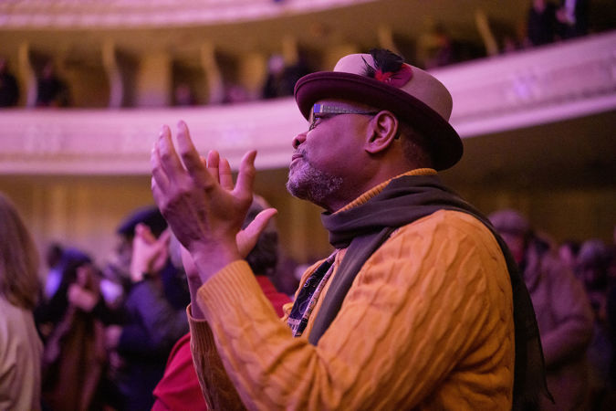 an audience member in a brown hat and orange sweater stands and claps after the performance