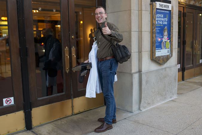 Principal Oboe William Welter, holding his concert wear, turns around and gives the camera a big smile and thumbs up as he enters the building