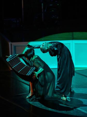 two women dressed in black dance on an eerie green backlit stage, while one plays the accordion-like bandoneon