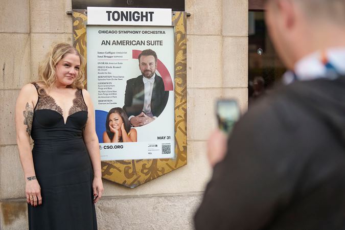 an audience member with blond, curly hair gets a photo taken next to the front of house poster on Michigan Avenue, all dressed up for the concert