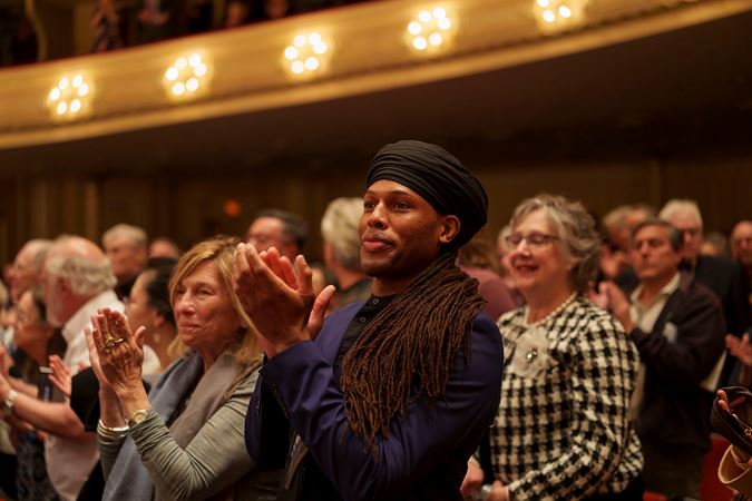an audience member on the main floor, in a blue shirt with long dark hair in a wrap, stands and claps