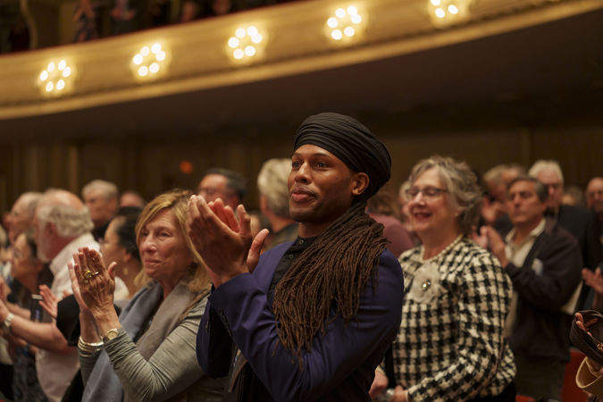 an audience member on the main floor, in a blue shirt with long dark hair in a wrap, stands and claps