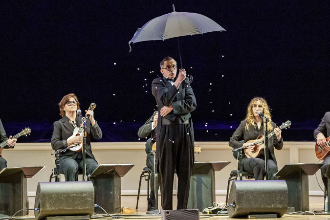 Guy Bellingham holds up an umbrella and shivers while fake snow drifts down around him on stage