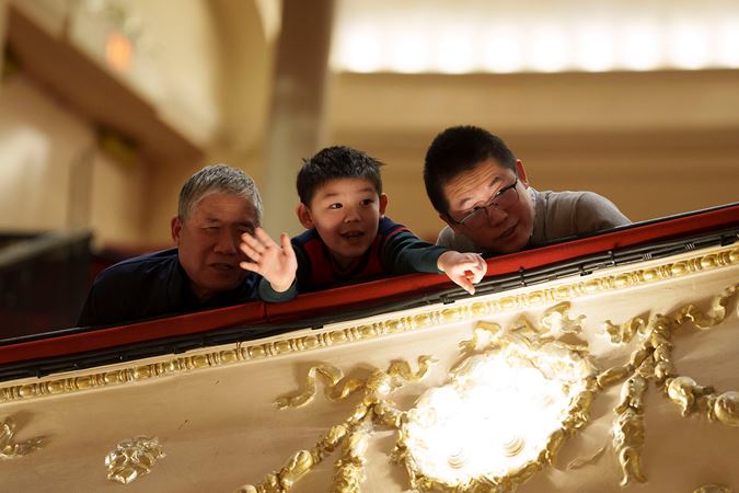 A young audience member pointing towards the stage and two adults looking from the Lower Balcony at the finalists on stage