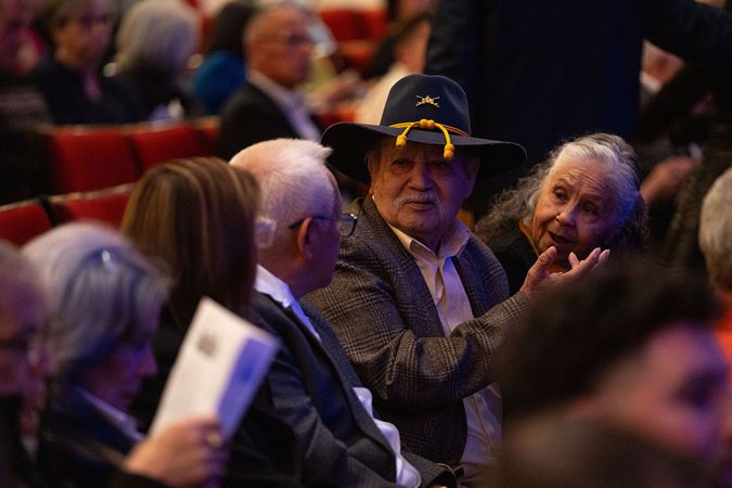 an audience member with a blue and yellow wide-brimmed hat and a mustache speaks animatedly to fellow concertgoers before the performance