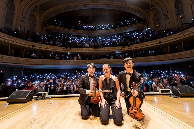 from left to right, violinist Brett Yang, pianist Sophie Druml, and violinist Eddy Chen pose on stage in front of a sea of phone flashlights