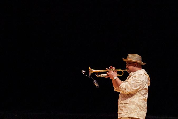 A side profile of Nicholas Payton performing on trumpet. He wears a tan jacket, a brimmed hat and glasses.