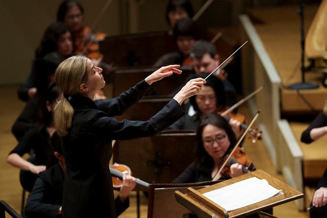 Conductor Karina Canellakis, with a baton in her right hand, lifts both arms to gesture to the full orchestra from the podium