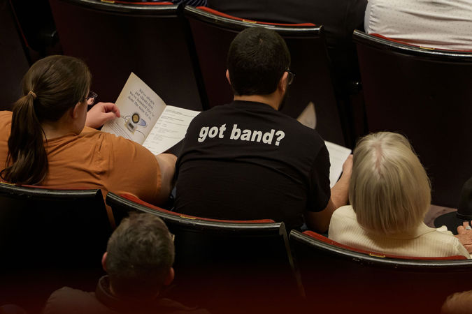 an audience member, wearing a black shirt with the slogan "got band?" on the back, reads their program