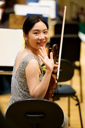 Closeup shot of CSO violin So Young Bae smiling and waving at the camera in a silver sequins gown