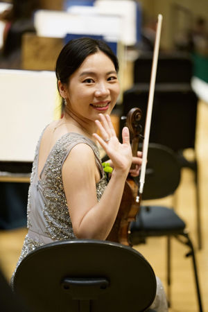 Closeup shot of CSO violin So Young Bae smiling and waving at the camera in a silver sequins gown