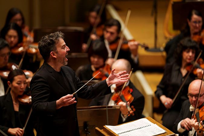 conductor James Gaffigan gives an enthusiastic, open-mouthed smile as he gestures to the Orchestra during a piece