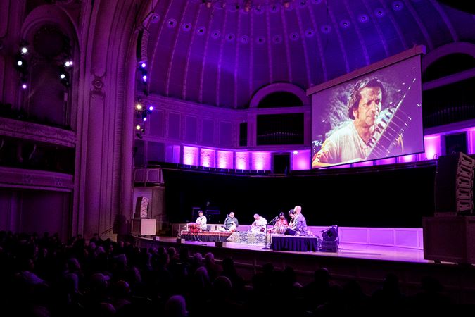 the Ravi Shankar Ensemble performs in front of a screen featuring an archival image of their namesake holding his sitar