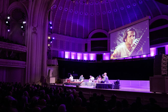 the Ravi Shankar Ensemble performs in front of a screen featuring an archival image of their namesake holding his sitar