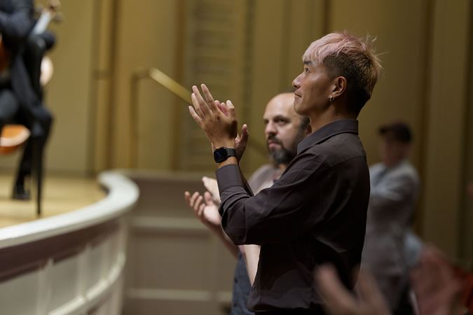 An audience member in the front row, with short pink hair and an elegant black watch, gives the orchestra a standing ovation
