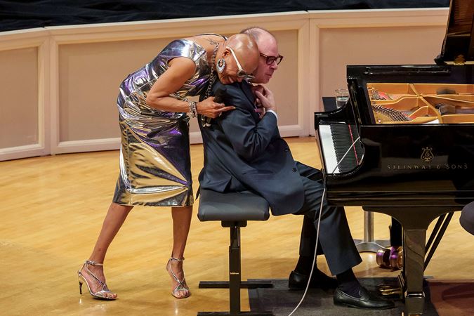 Dee Dee Bridgewater, wearing a sparkly silver dress and heels, hugs her collaborator, Bill Charlap, from behind as he sits at the piano