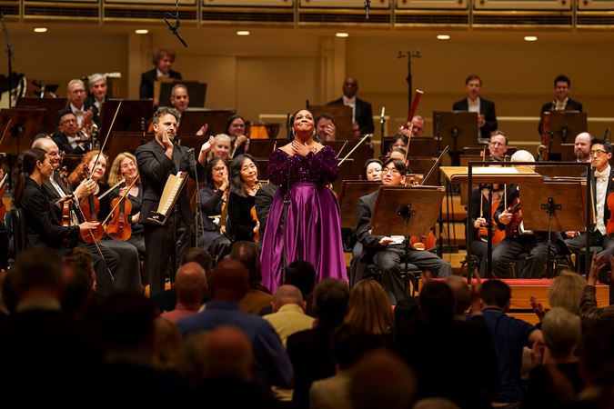 Soprano Janai Brugger, wearing a purple dress on stage, clasps her hands in gratitude as she bows before the audience