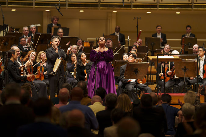 Soprano Janai Brugger, wearing a purple dress on stage, clasps her hands in gratitude as she bows before the audience