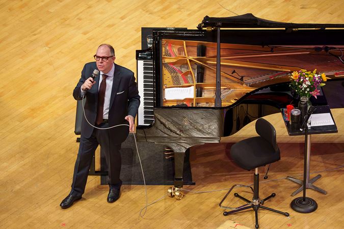 Bill Charlap stands next to the piano in a suit and addresses the audience