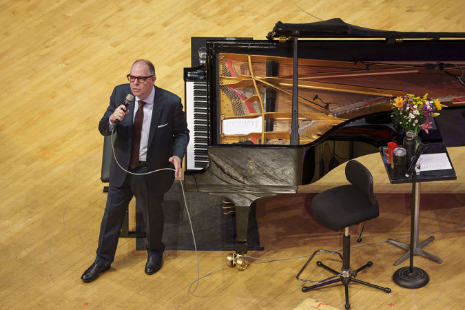 Bill Charlap stands next to the piano in a suit and addresses the audience