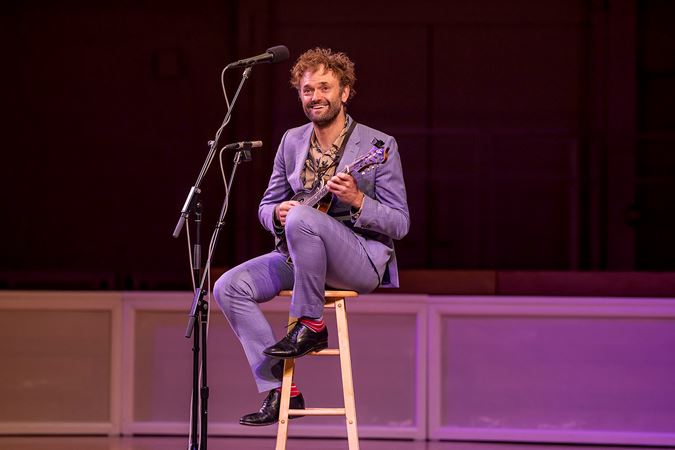 Chris Thile sits on a stool in a periwinkle suit, mandolin in hand and smiling out at the audience