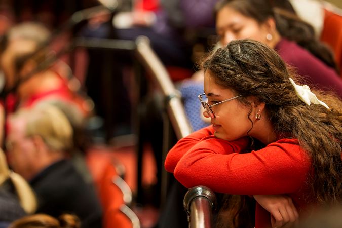 a young audience member in a red sweater and glasses looks down from the balcony
