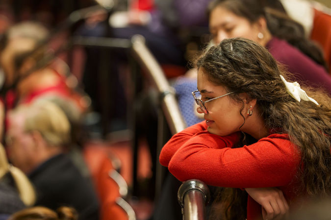 a young audience member in a red sweater and glasses looks down from the balcony