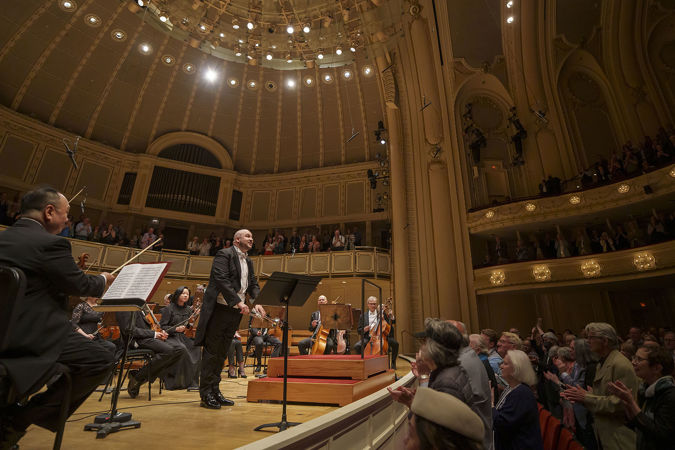 Side wide shot of CSO principal trumpet Esteban Batallán bowing on stage and the audience applauding