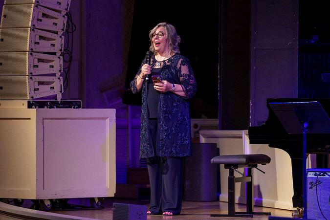 Kelly Peterson, Oscar Peterson's widow, stands on stage in a navy blue outfit and addresses the crowd