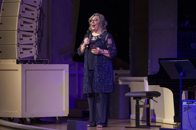 Kelly Peterson, Oscar Peterson's widow, stands on stage in a navy blue outfit and addresses the crowd