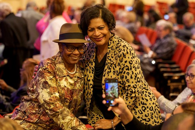 two fans, dressed in tan blouses and stylish jewelry, pose near their seats for a phone photo before the concert