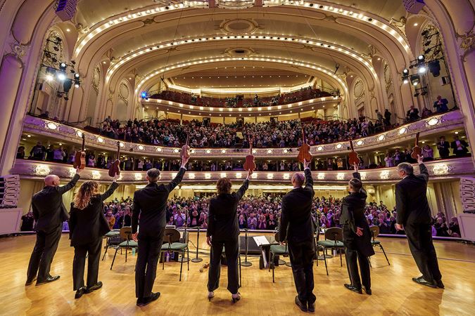 point of view from the stage behind the Ukulele Orchestra of Great Britain members, as they raise their instruments in the air in preparation for a bow