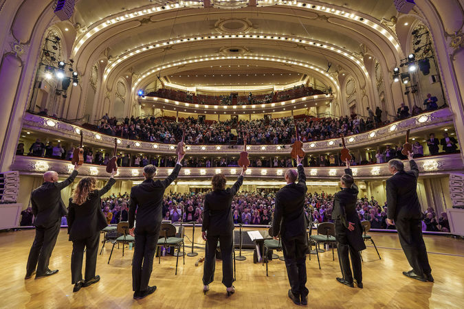 point of view from the stage behind the Ukulele Orchestra of Great Britain members, as they raise their instruments in the air in preparation for a bow