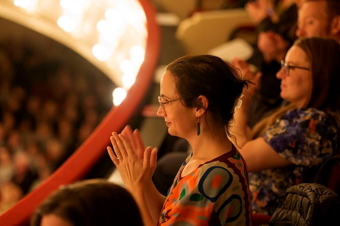 an audience member in glasses and a colorful blouse looks on and applauds from the box level