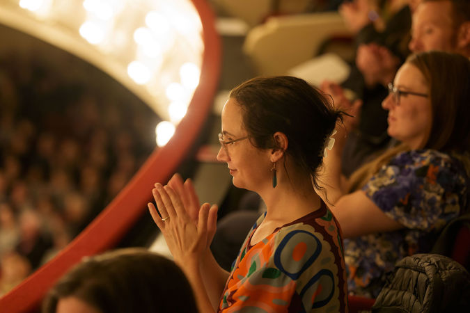 an audience member in glasses and a colorful blouse looks on and applauds from the box level