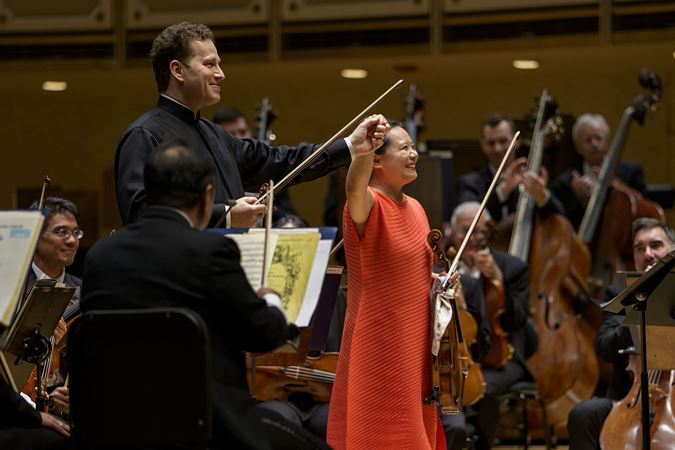 Nikolaj Szeps-Znaider and Teng Li raise their hands in a bow. Szeps-Znaider wears a black dress shirt and Li an orange pantsuit.
