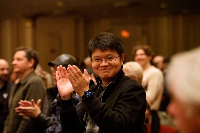 an audience member in glasses and a dark jacket, sitting in the front row, smiles at the camera as he claps for the CSO