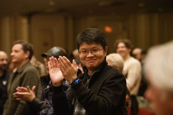 an audience member in glasses and a dark jacket, sitting in the front row, smiles at the camera as he claps for the CSO