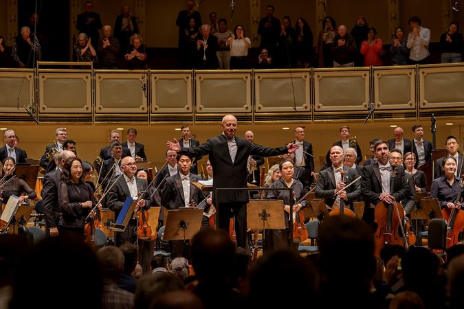 on the podium, Paavo Järvi spreads his arms in an invitation for the full orchestra to bow