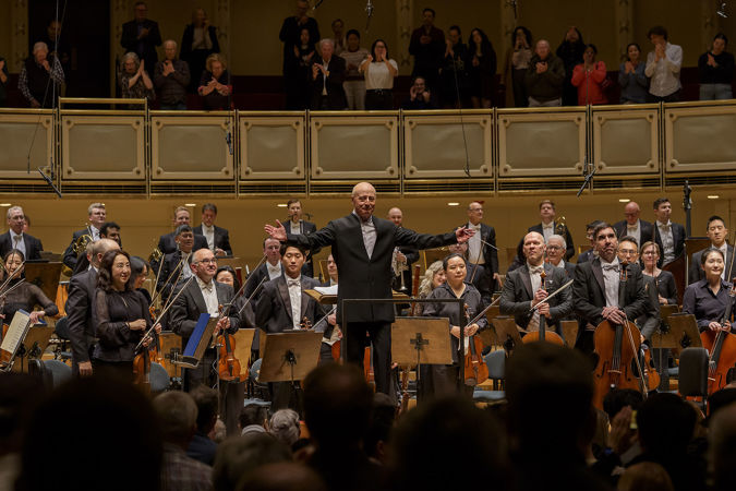 on the podium, Paavo Järvi spreads his arms in an invitation for the full orchestra to bow